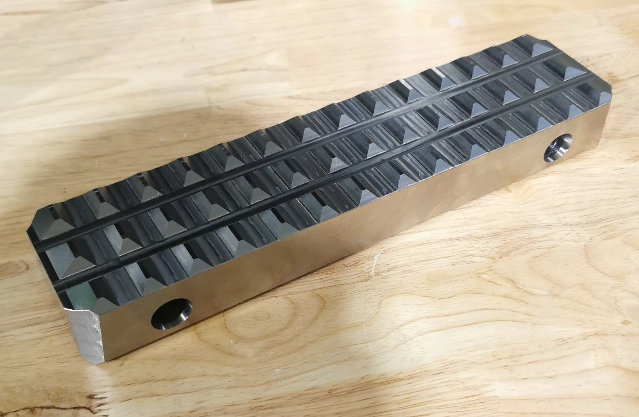 A metal linear rail with evenly spaced triangular ridges on the top surface, placed on a wooden surface.