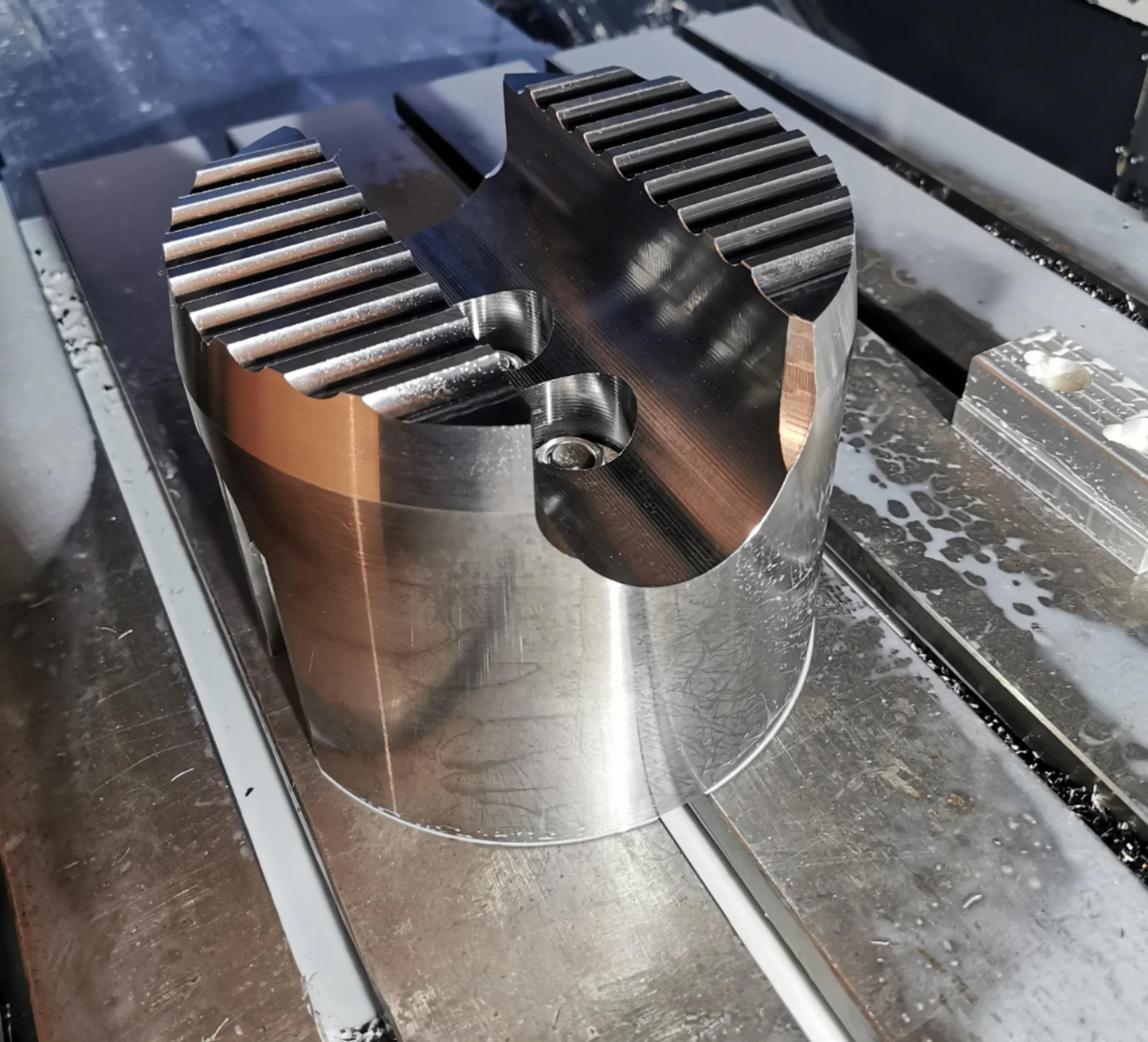 A metal machined component with gear-like teeth on the top edge, sitting on a worktable with metal shavings and machining tools.