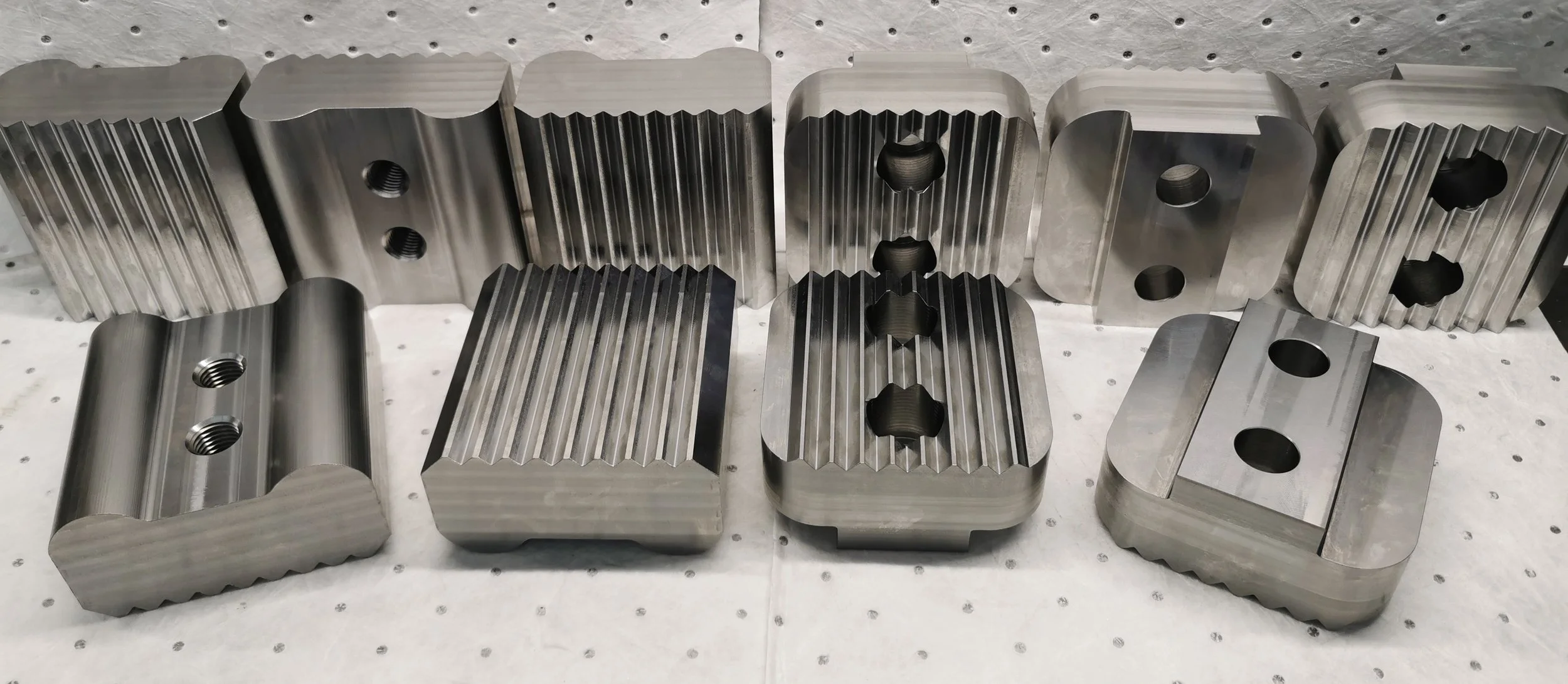 Several machined metal parts with beveled edges and threaded holes, arranged in a row on a perforated work surface.
