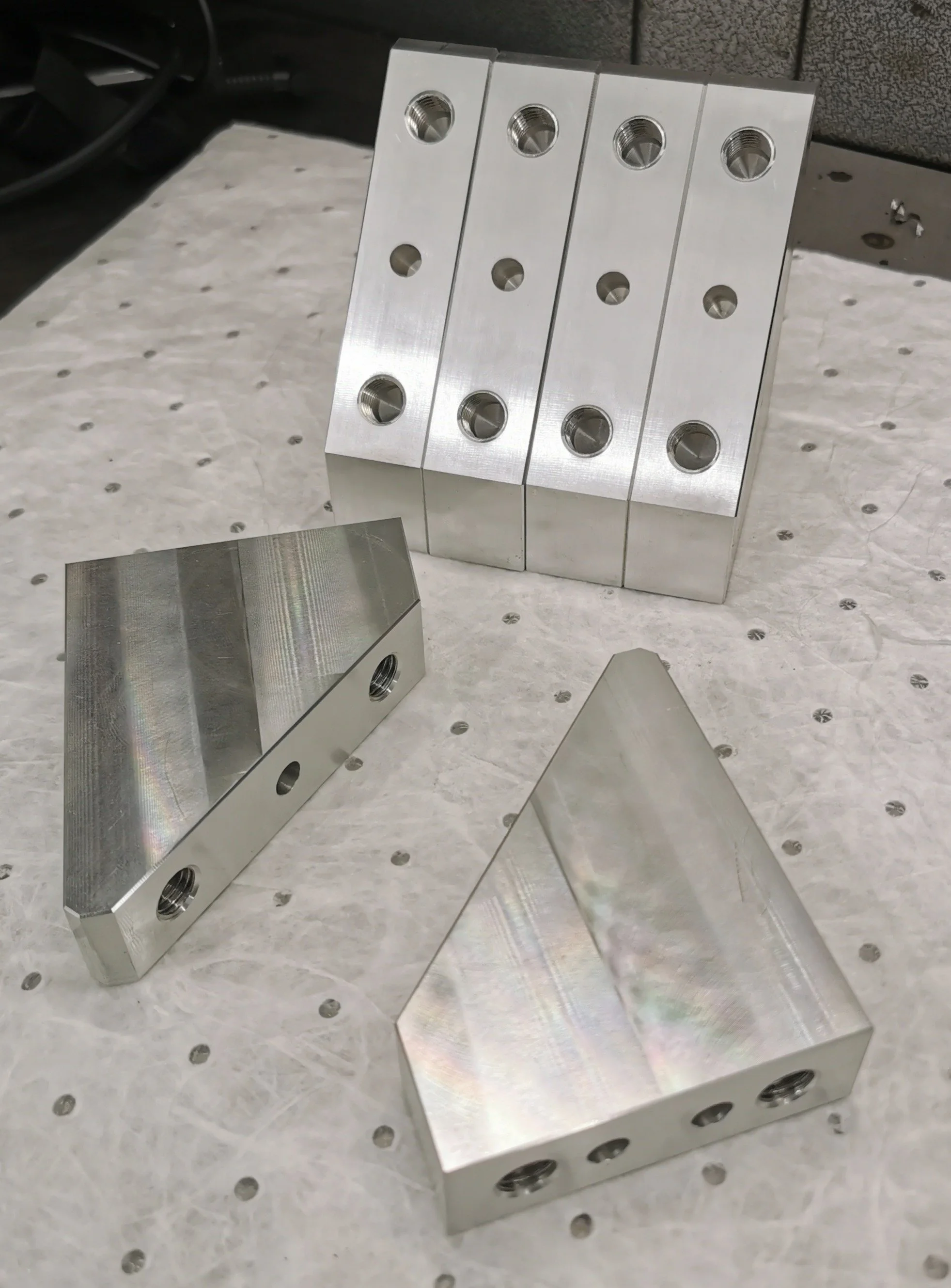 Several machined metal blocks with threaded holes placed on a perforated work surface, likely to be used for manufacturing or assembly.