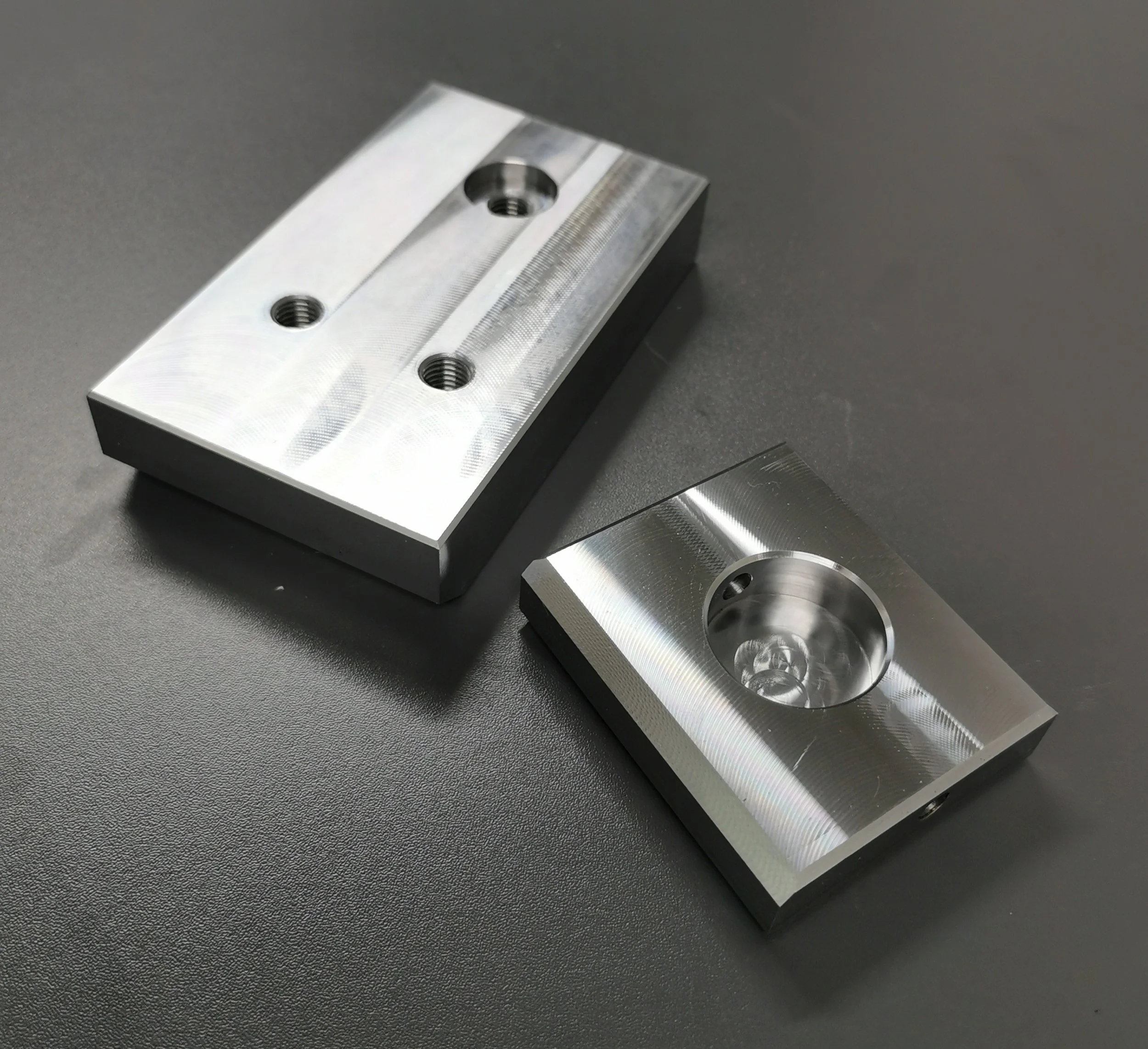 Two machined metal blocks with threaded holes, placed on a black surface.