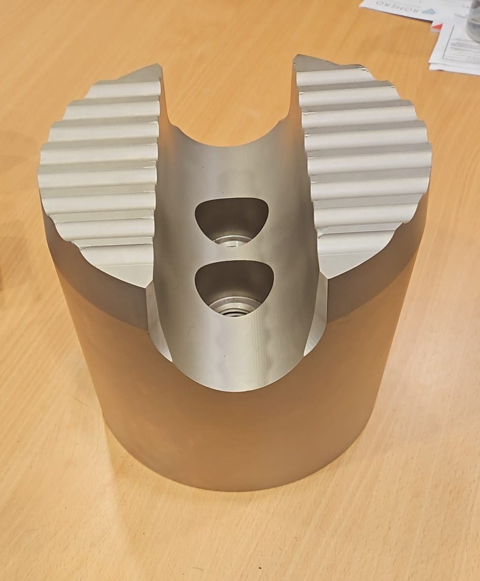 Metal machine part with two large holes and ribbed sections on the upper sides, placed on a wooden surface, with papers and a glass in the background.