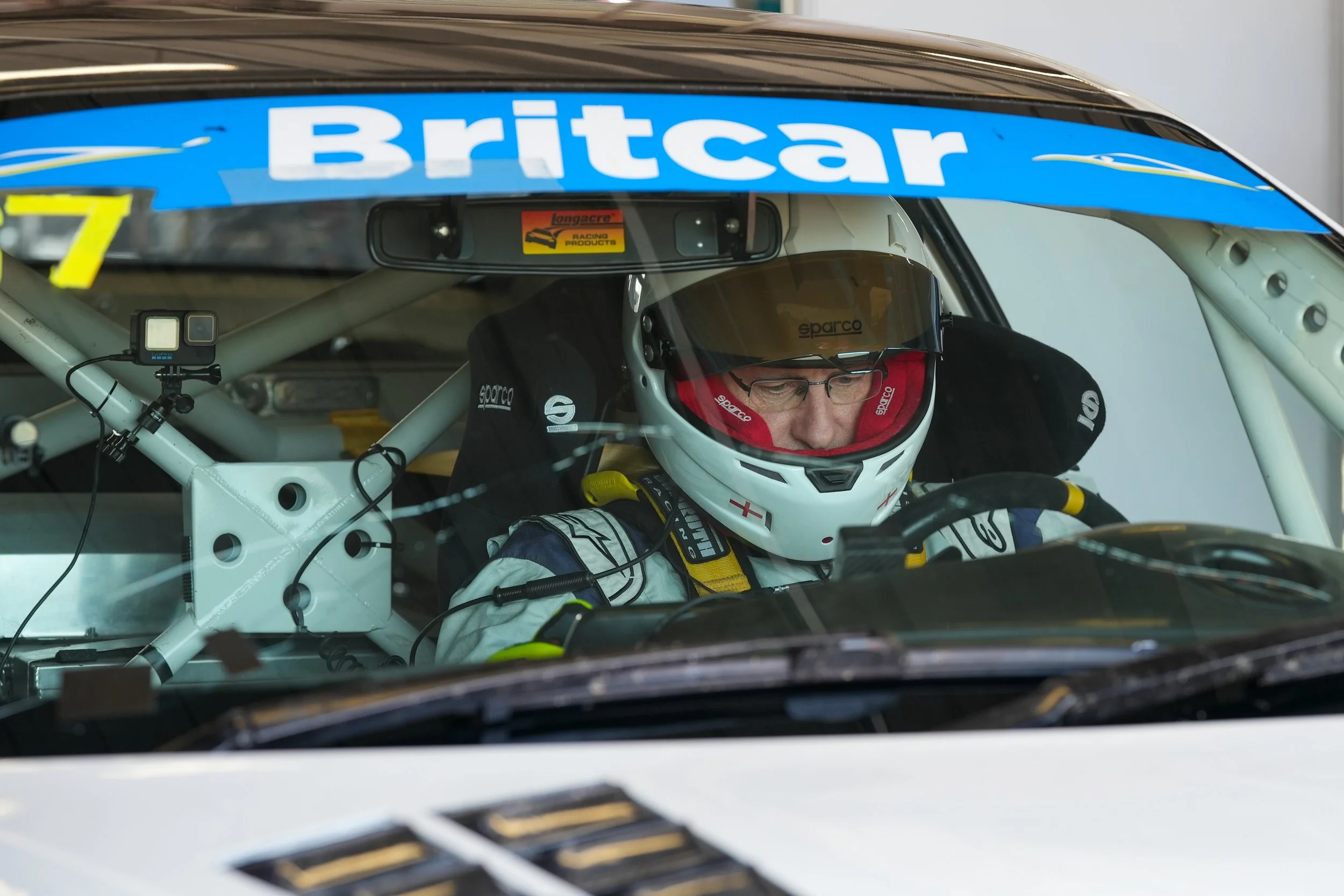 A race car driver wearing a helmet and racing suit inside the car, with a blue banner with the word 'Britcar' hanging above the windshield.