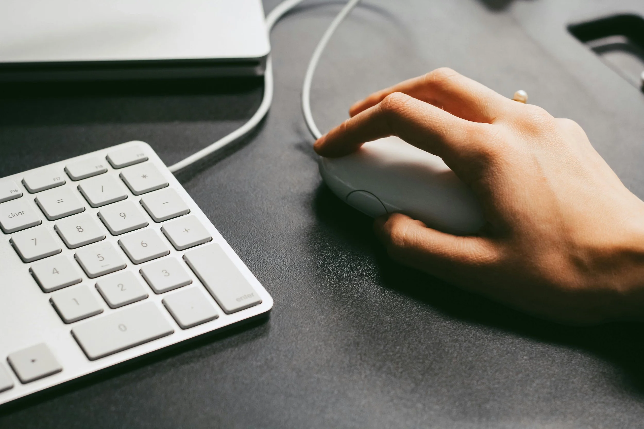 A person's hand using a computer mouse on a desk with a white keyboard nearby.