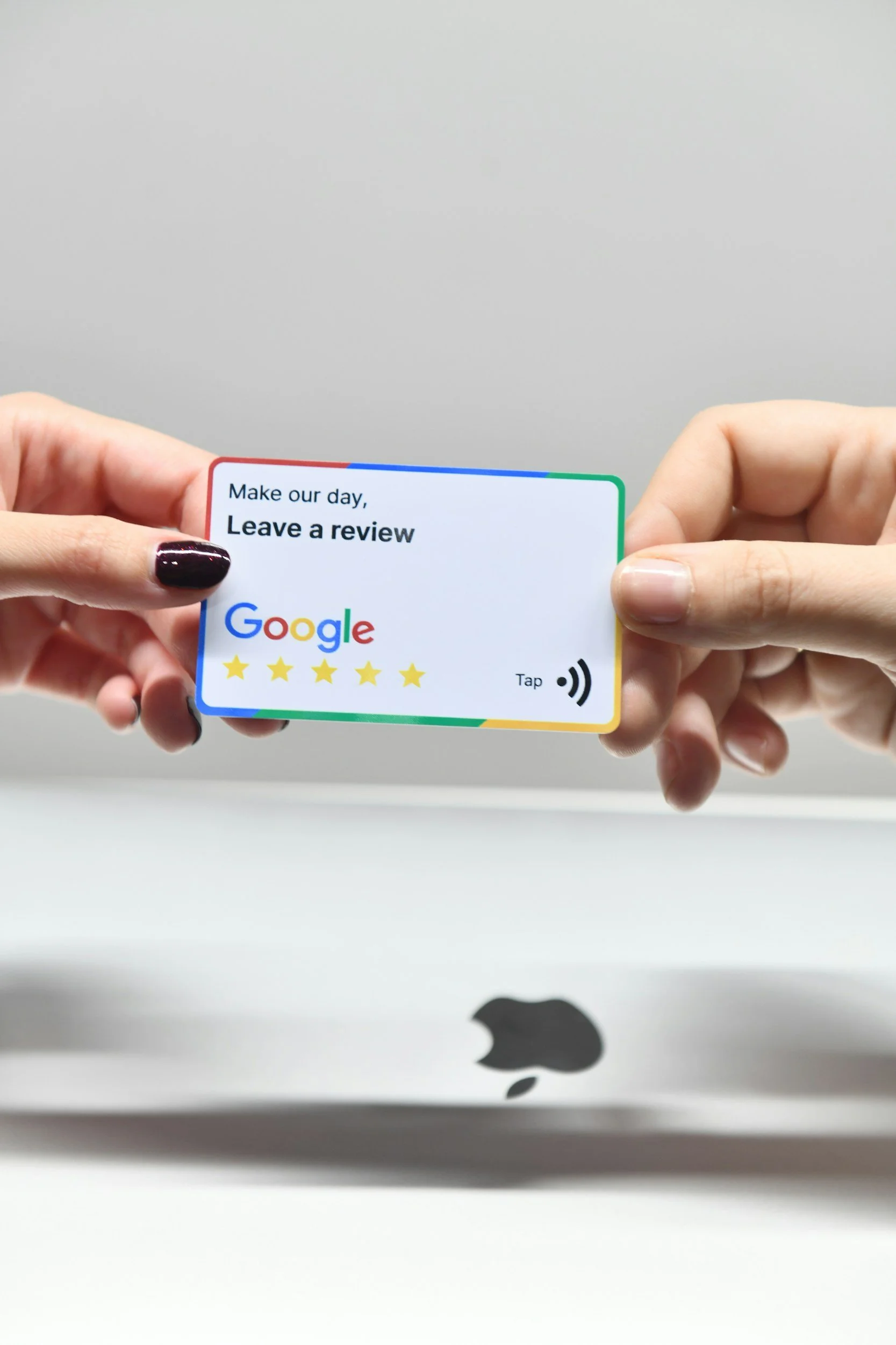 A person holding a colorful Google review card in front of a silver MacBook with an Apple logo, against a plain gray wall background.