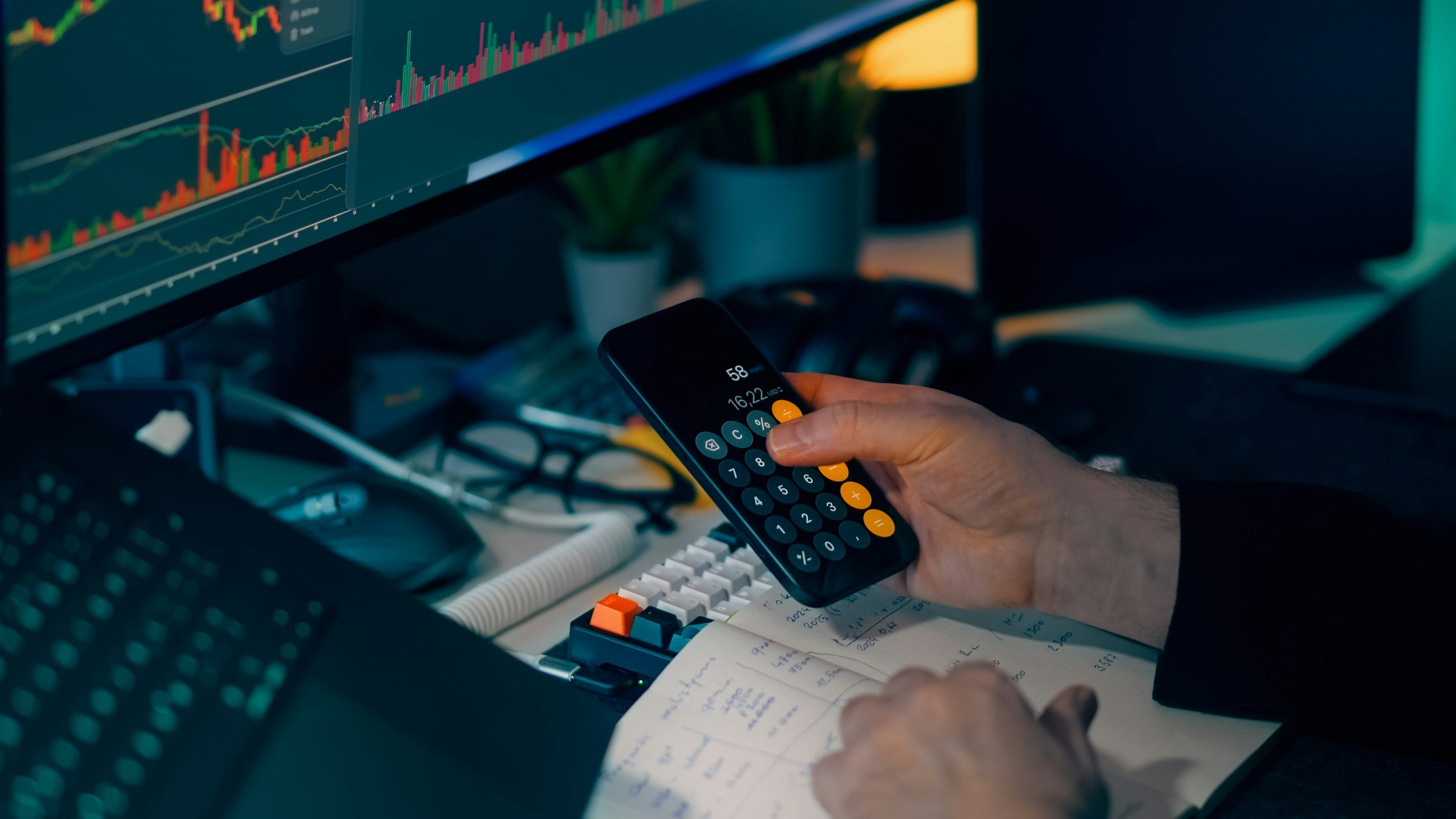 A person holding a calculator in front of a computer monitor displaying financial charts with candlestick graphs, surrounded by office items like a keyboard, notepad with notes, and a desk with various electronic devices.