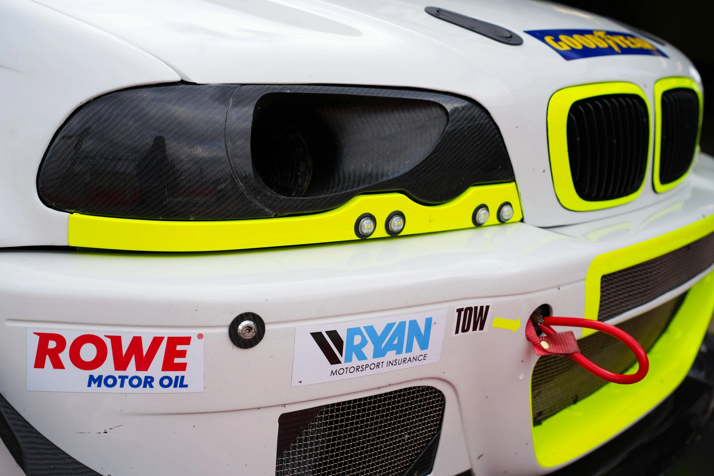 Close-up of a race car's front end showing black carbon fiber headlights, a white body with team and sponsor stickers, and yellow accents around the grille. Red wires are visible near the lower right corner.