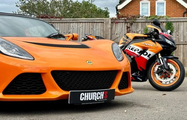 An orange sports car parked next to a black, orange, and white Repsol Honda racing motorcycle in a residential driveway with a wooden fence and brick house in the background.