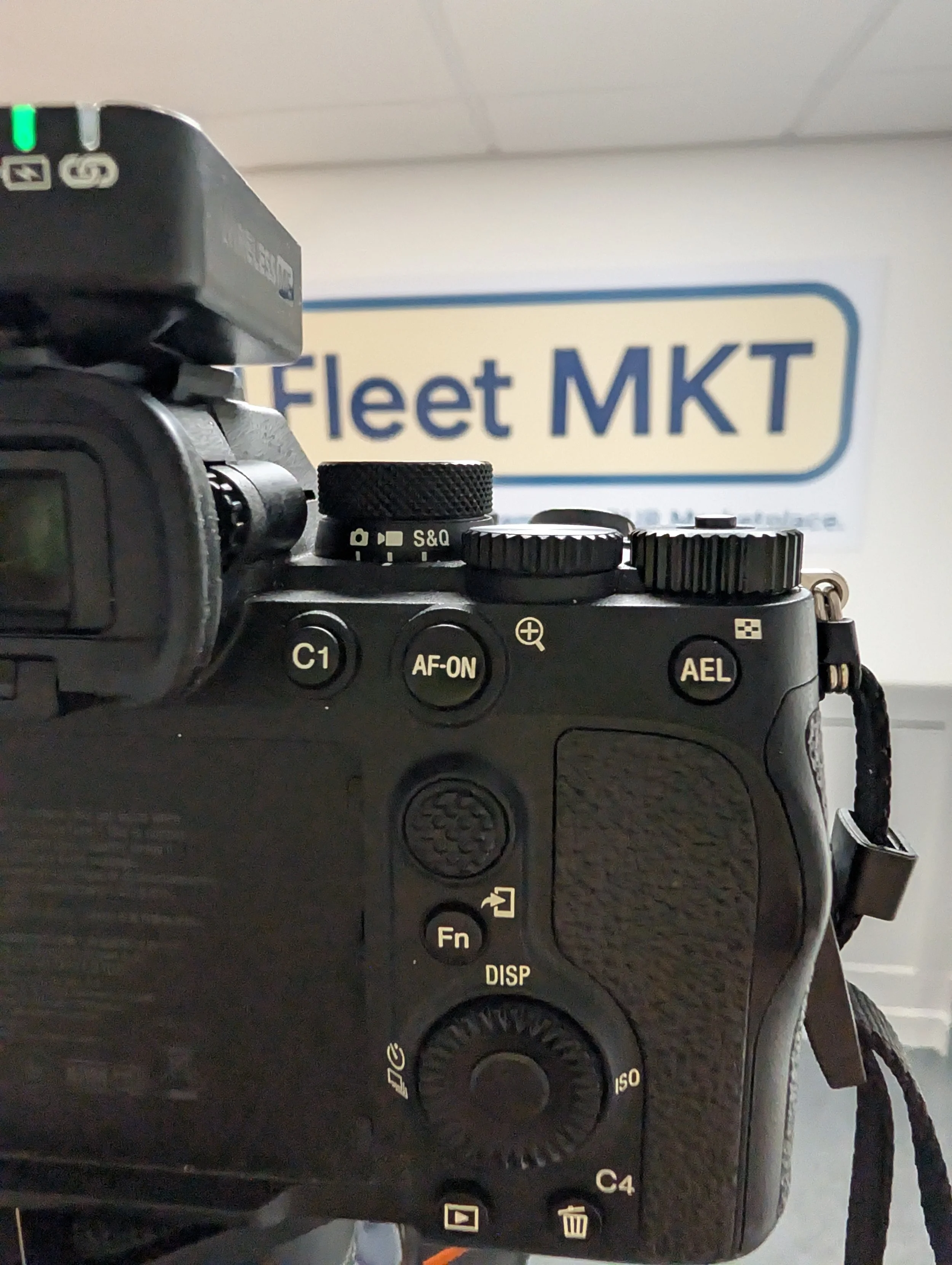 Close-up of a black digital camera showing various buttons and a control dial, with a sign in the background reading 'Fleet MKT' in blue text on a white background.