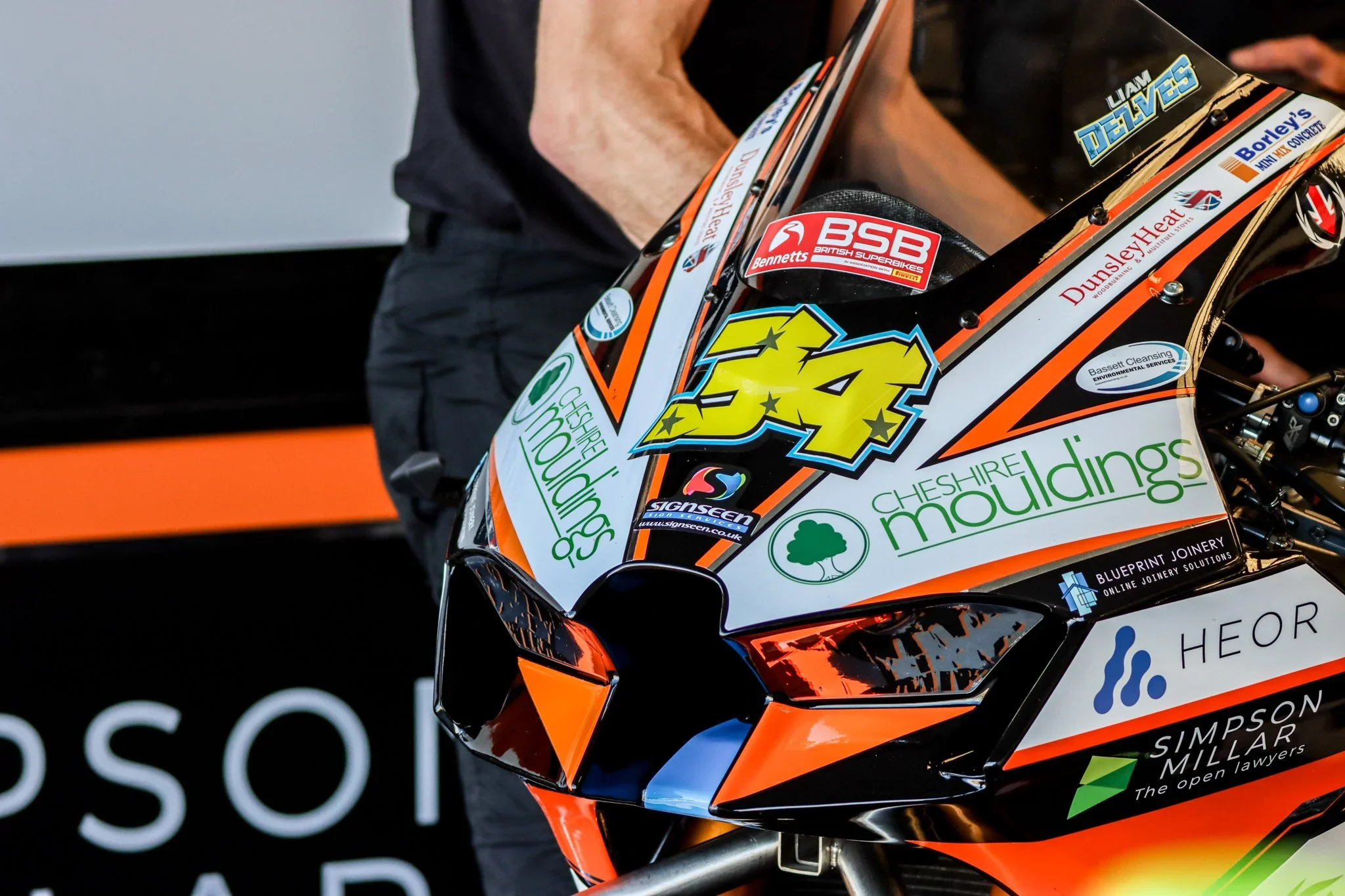 Close-up of a racing motorcycle with sponsors' stickers, including the number 43, and a person wearing black in the background.