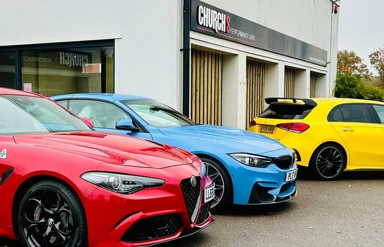 Three cars parked outside a building with a sign that reads 'CHURCHES' and 'EXPERIENCE CARS.' The cars are red, blue, and yellow, with the red and blue cars appearing sports models, and the yellow car a hatchback.
