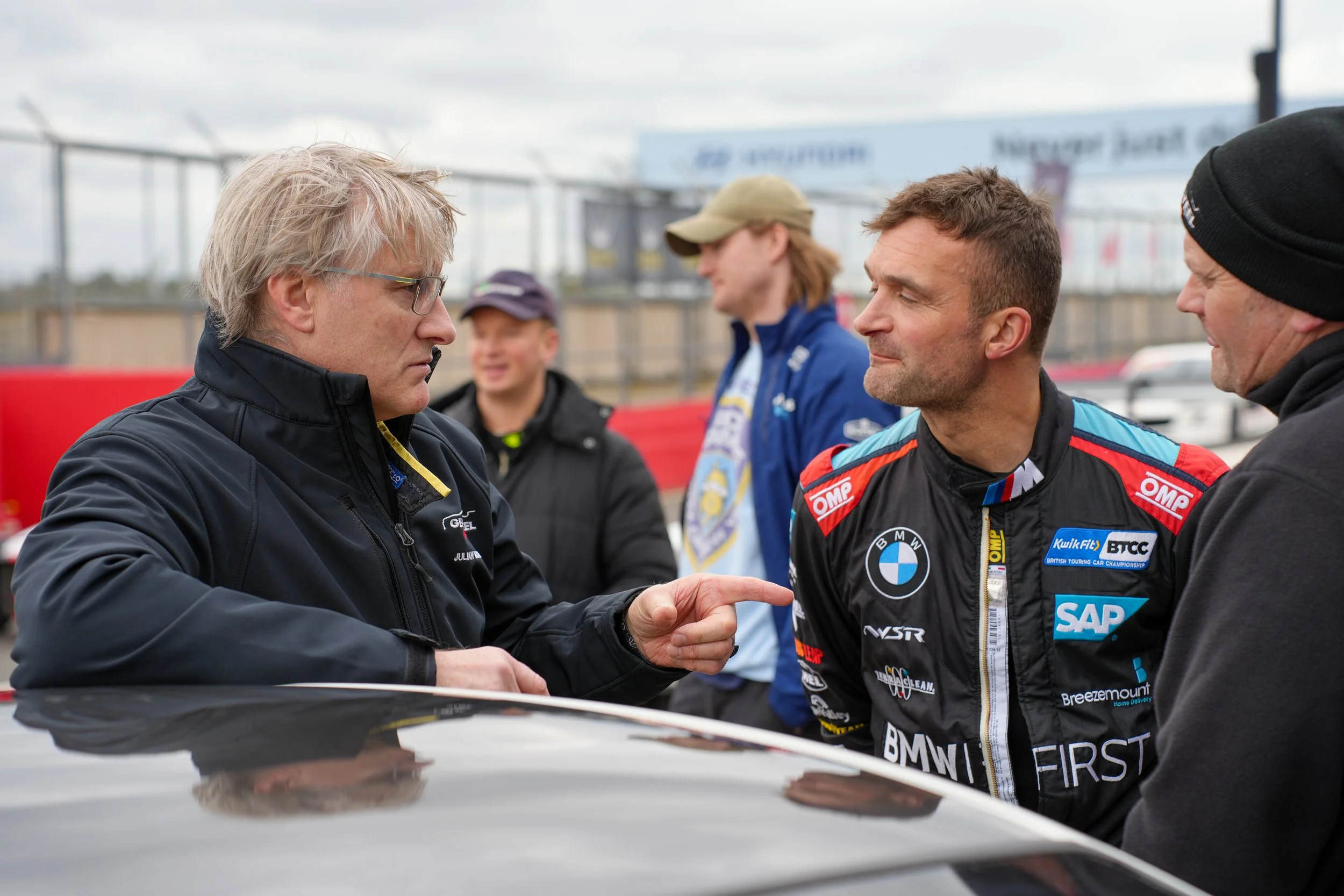 Two men shaking hands while others look on at a race track, with a background of a cloudy sky and a fence.