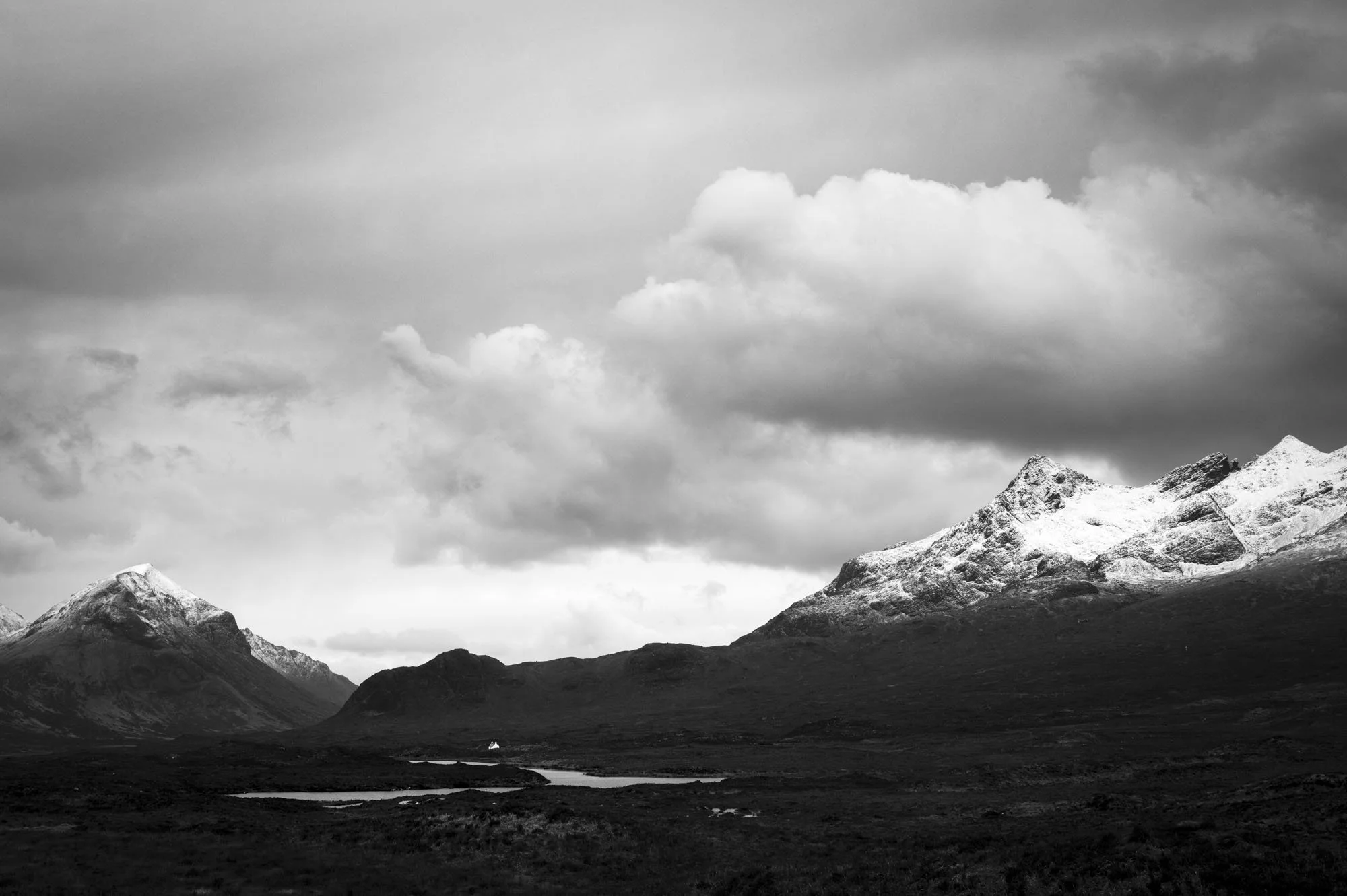 Black and white photograph of snow-capped mountains under cloudy sky with a small lake in the foreground.
