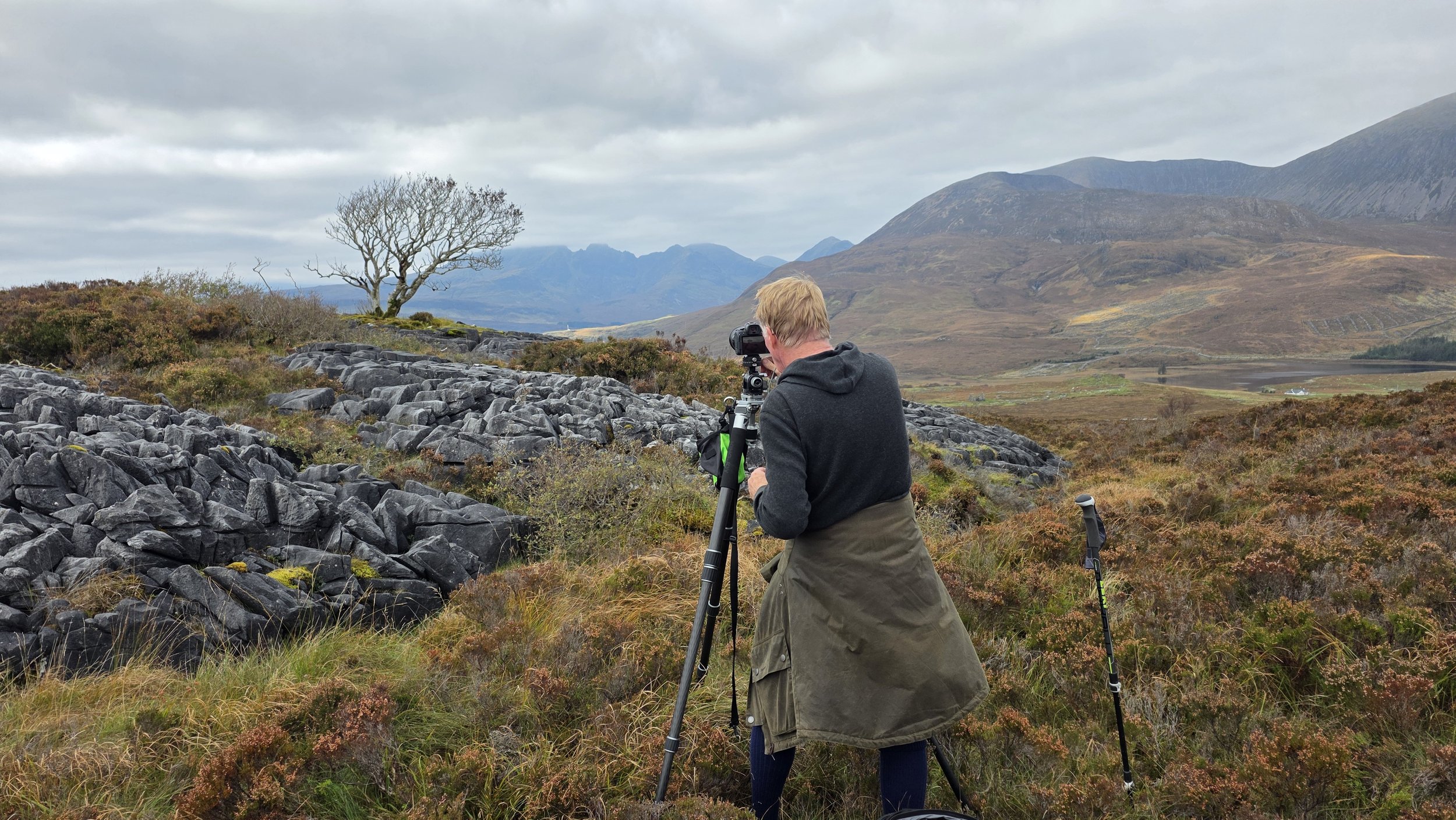 Two photographers on a Lens57 photography workshop with tripods and cameras set up on rocky hillside of the Isle of Skye in a mountainous landscape with leafless tree in the distance.