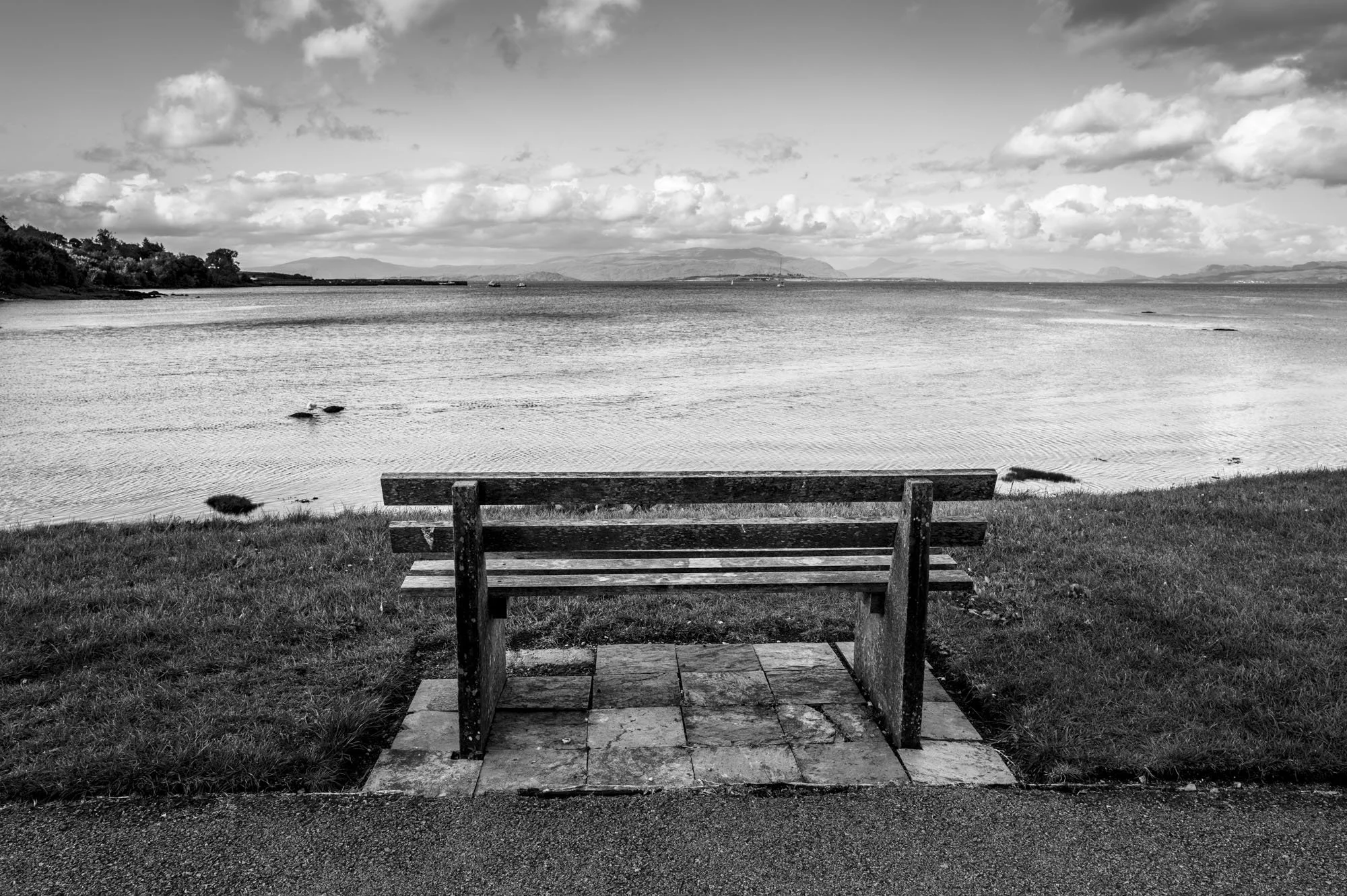 A black and white photo of a wooden bench on a paved area facing a calm body of water with hills in the background and a partly cloudy sky.