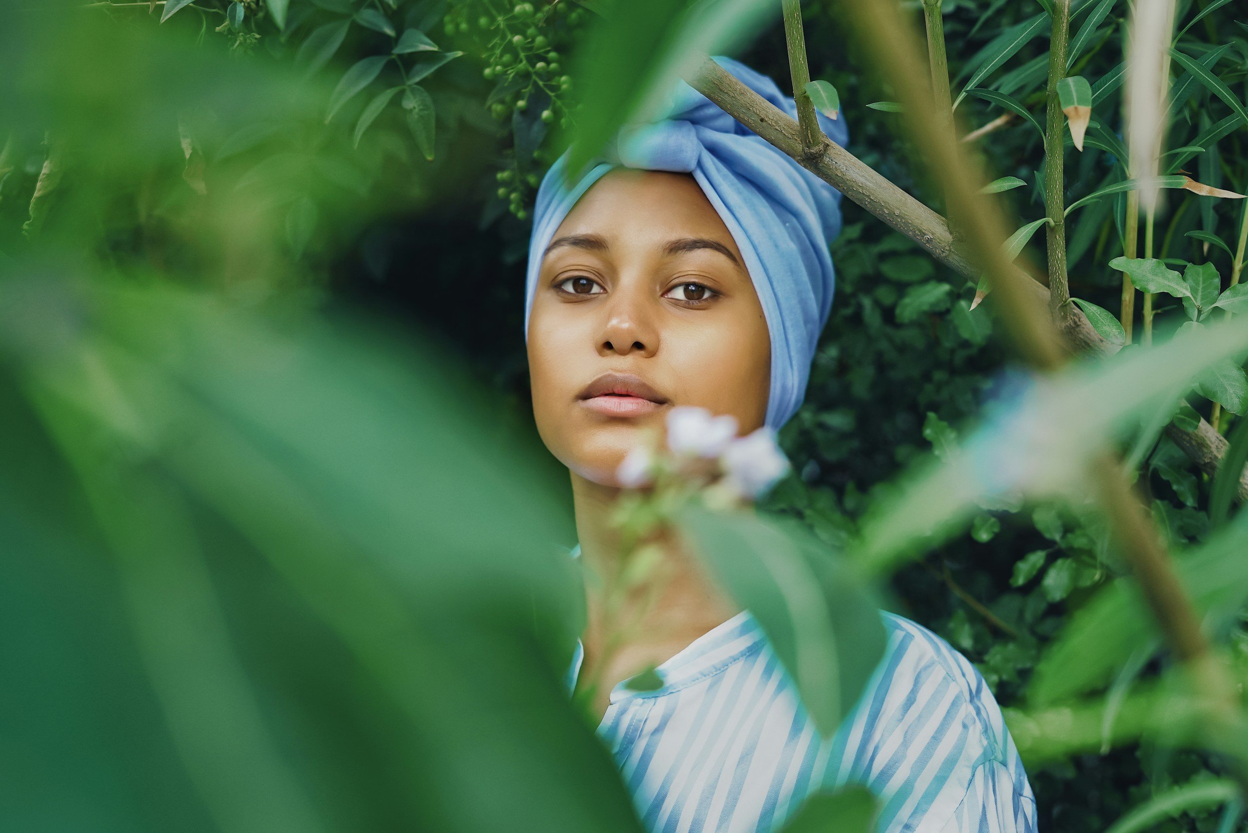 A woman wearing a light blue headwrap and striped shirt is partially visible through green foliage.