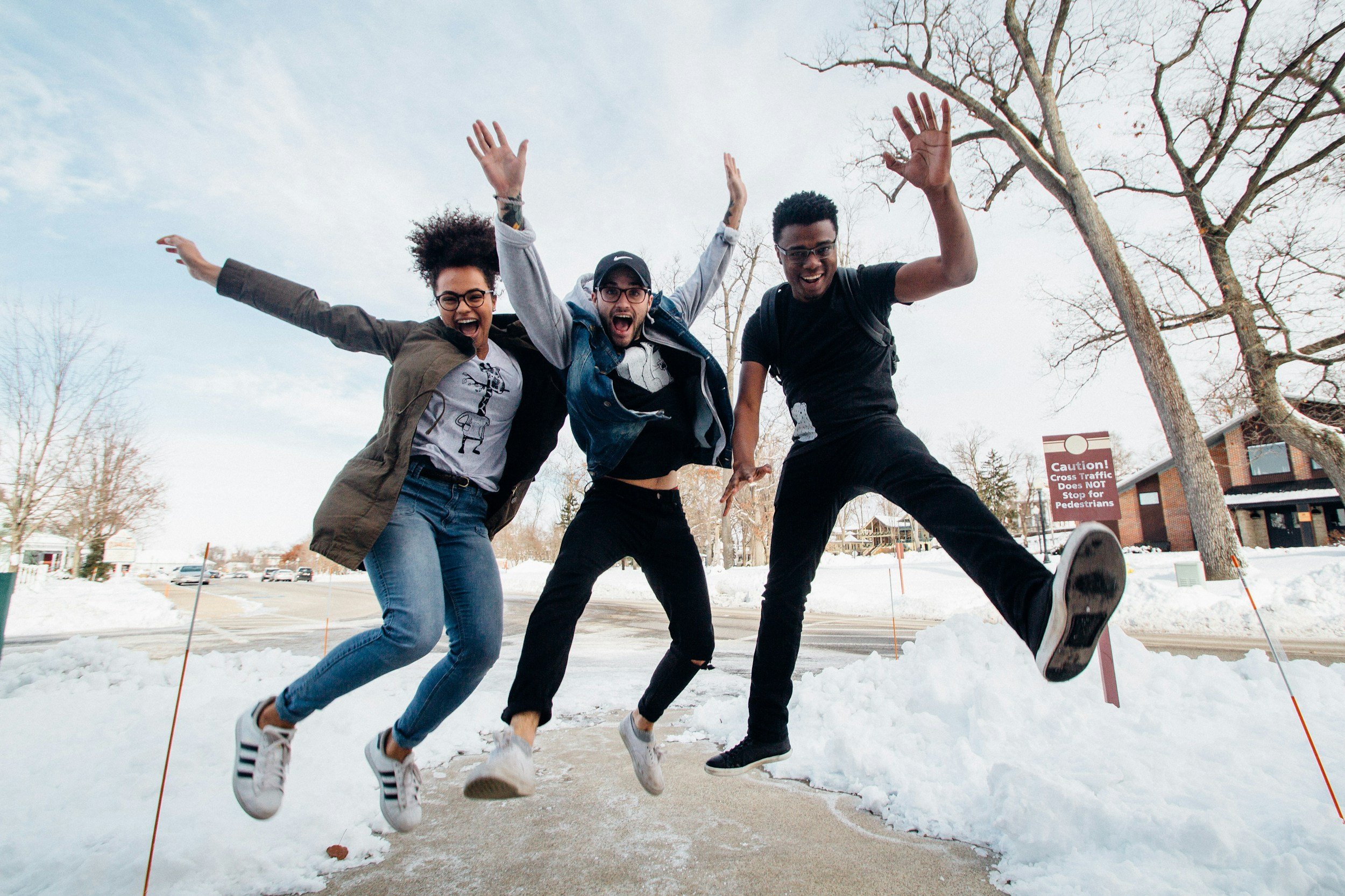 Diverse group of three people jumping with joy on a snowy street, wearing casual winter clothing, with trees and a caution sign in the background.
