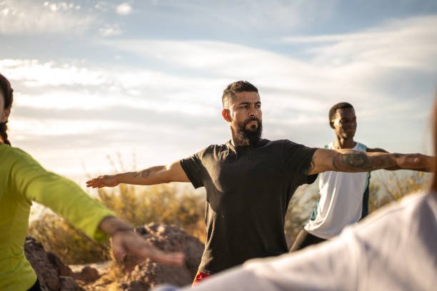 Group of diverse people practicing yoga outdoors, standing in a warrior pose with arms extended under a sunny sky.