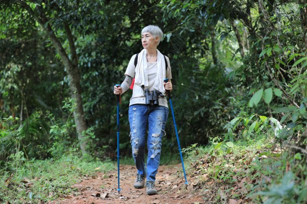 An older adult with gray hair hiking on a forest trail, using trekking poles and carrying binoculars.