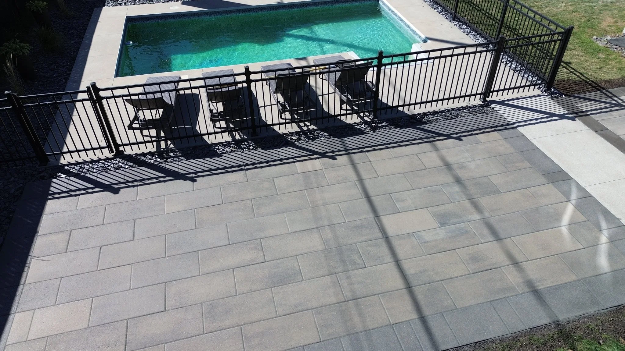 Piscine rectangulaire entourée d'une clôture noire, avec plusieurs chaises longues sur le patio en pavés, et ombres projetées par la clôture.