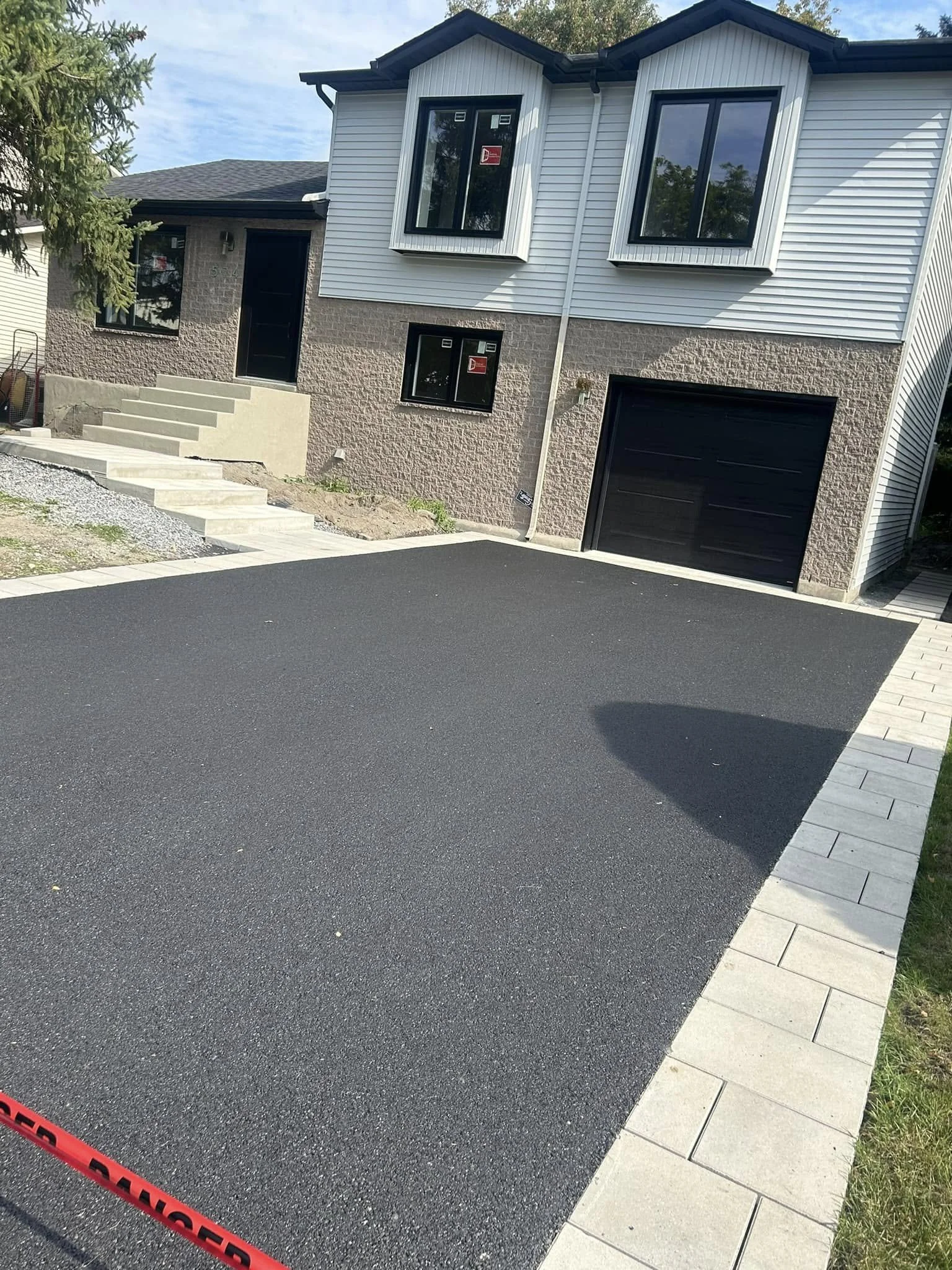Nouvelle allée asphaltée devant une maison moderne avec garage noir, fenêtres grandes et porte d'entrée en hauteur, avec des escaliers menant à l'entrée principale.