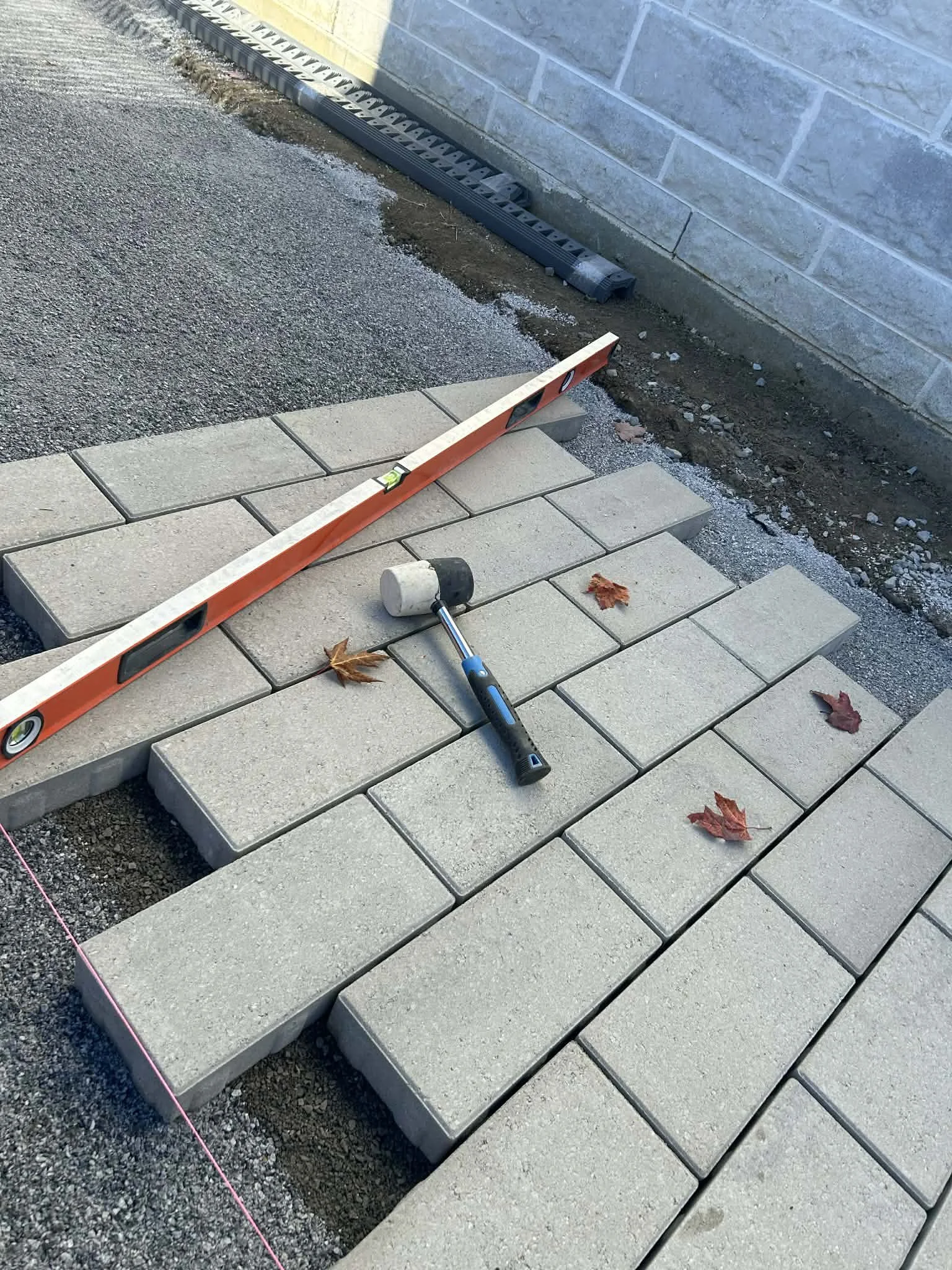 Pose de pavés en pierre avec niveau à bulle, marteau, taloche en plastique, et feuilles d'automne autour, sur un chantier de construction ou de rénovation d'une allée ou d'une terrasse.