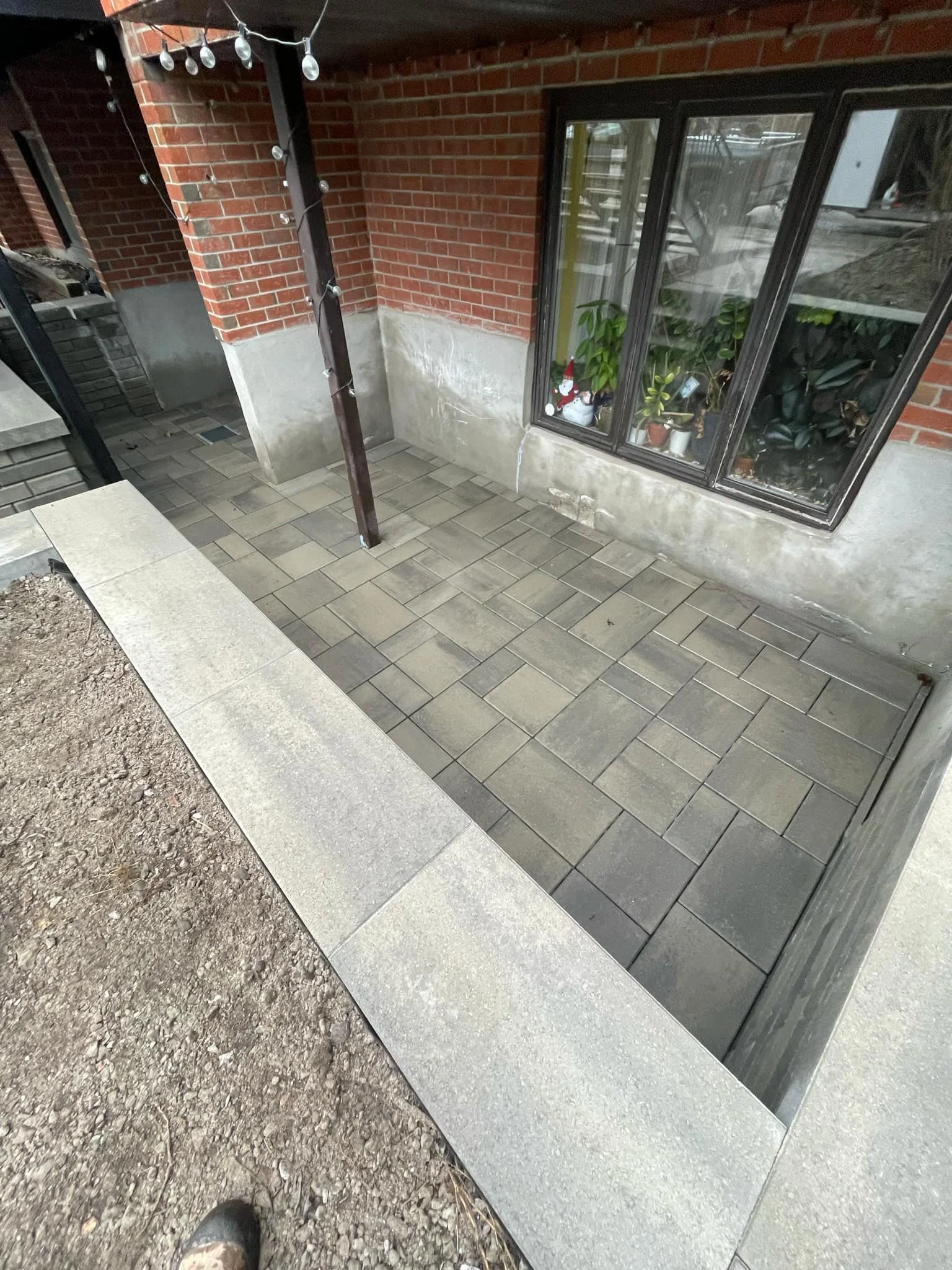 Un petit patio ou balcon avec un sol en carreaux gris, entouré d'une bordure en pierre, avec une fenêtre à cadre noir et quelques plantes à l'intérieur, et une maison en brique rouge en arrière-plan.