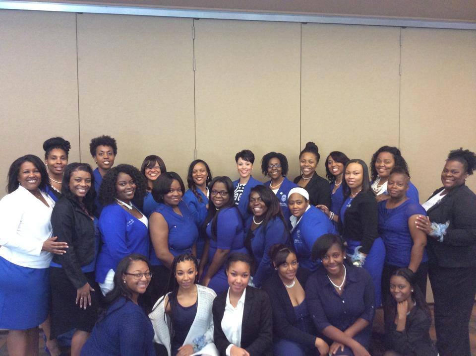Group of women in blue and black business attire posing for a photo in front of a plain beige wall.