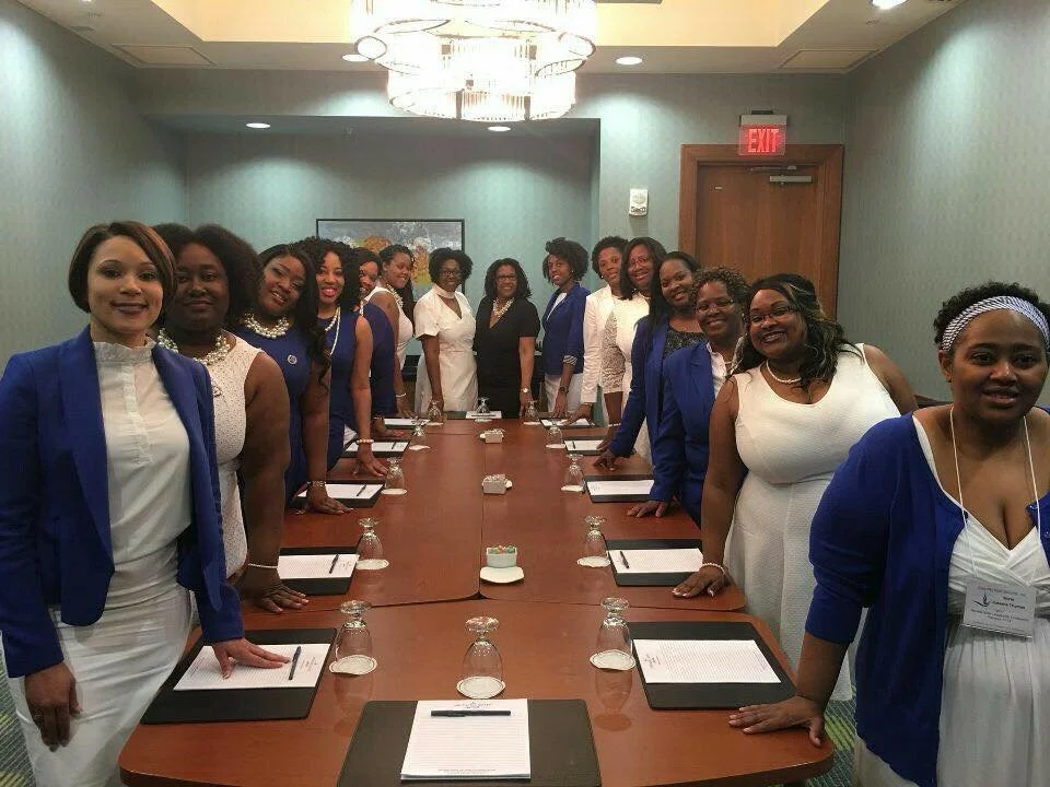 A group of women dressed in business attire, some in white and others in blue blazers, standing around a conference table in a meeting room, smiling for the photo.