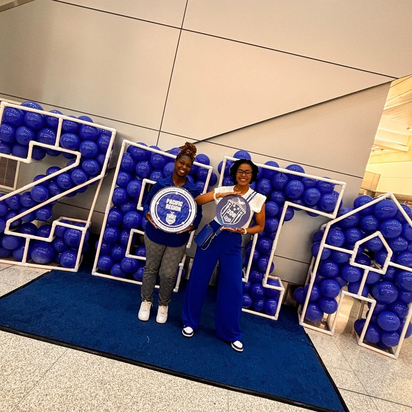 Two women standing in front of large blue number decorations with golf balls, holding round signs that read "Pacific Region" and "Zone 4" at an indoor event.