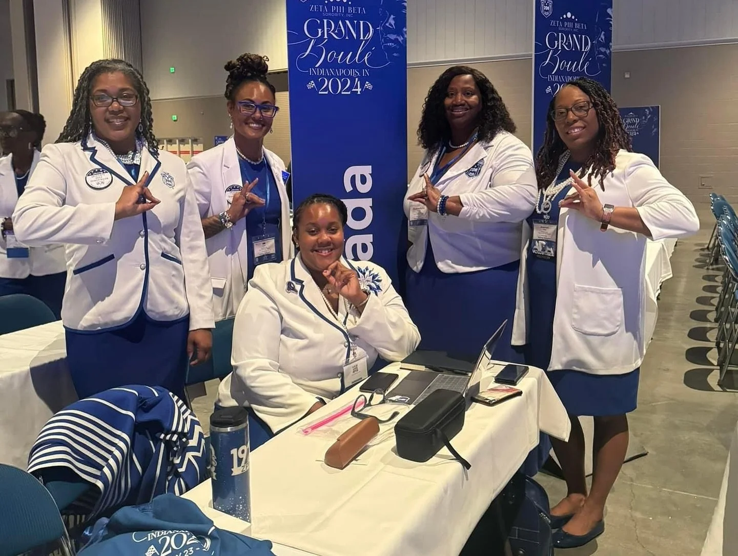 Six women dressed in white and blue uniforms, posing at a conference. One woman is seated at a table with a laptop, and the others are standing behind her, all making a peace sign. There are blue banners in the background with the text "Grand Boule, 