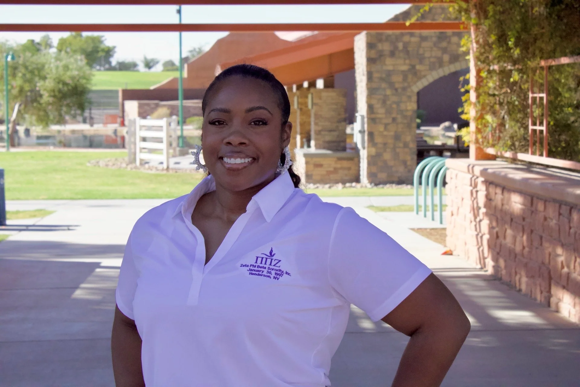 Smiling woman wearing a white polo shirt with purple embroidery, standing outdoors in a park or plaza area with buildings and trees in the background.