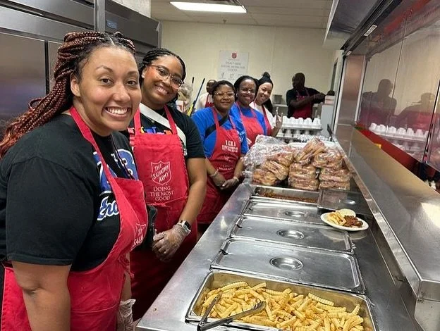 Group of smiling women wearing red aprons and black shirts serving food in a kitchen or cafeteria. There are trays of fried food and packaged bread.