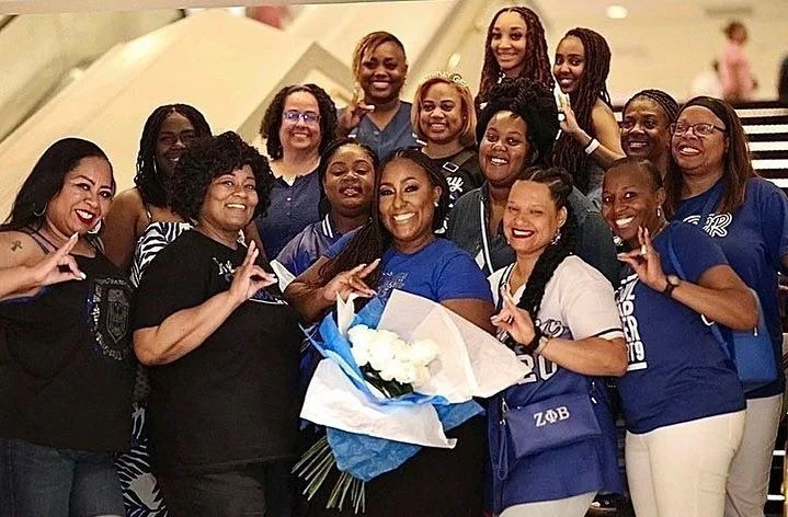 A group of women posing happily together, some holding a bouquet of white flowers. They are in an indoor setting with stairs visible in the background, wearing casual and team apparel.