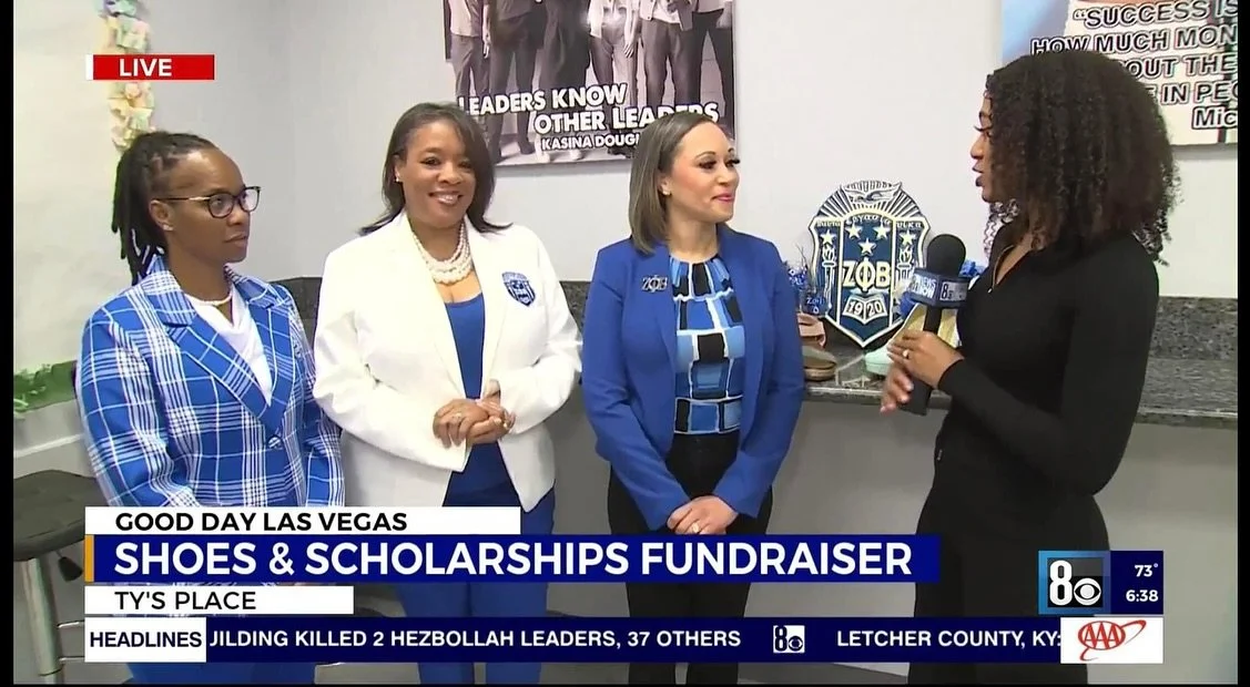 Four women standing indoors during a news interview, with a woman holding a microphone on the right. The women are dressed in blue and white outfits, and a banner in the background reads 'Leaders Know Other Leaders'.