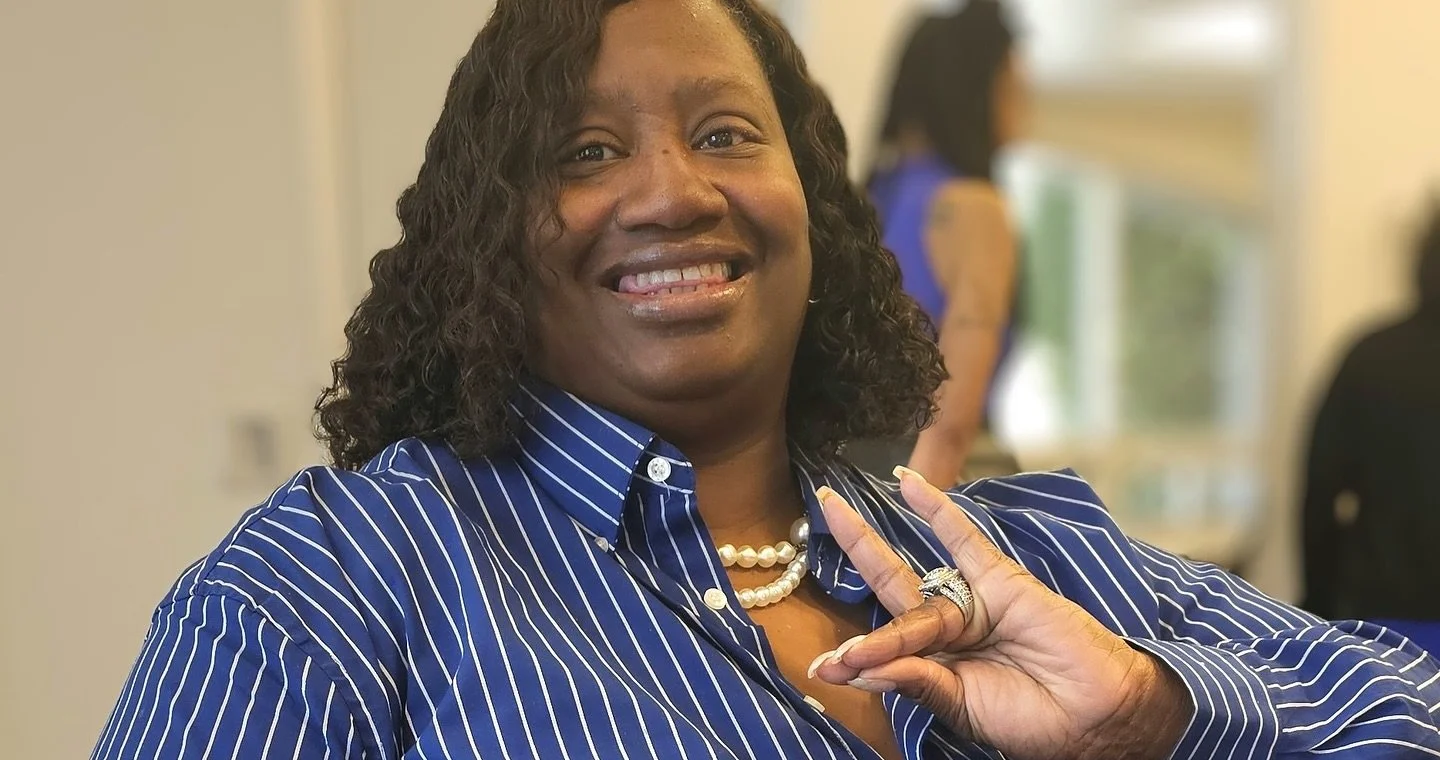 A smiling woman with dark curly hair wearing a blue and white striped shirt, pearl necklace, and rings, holding up a peace sign with her fingers.