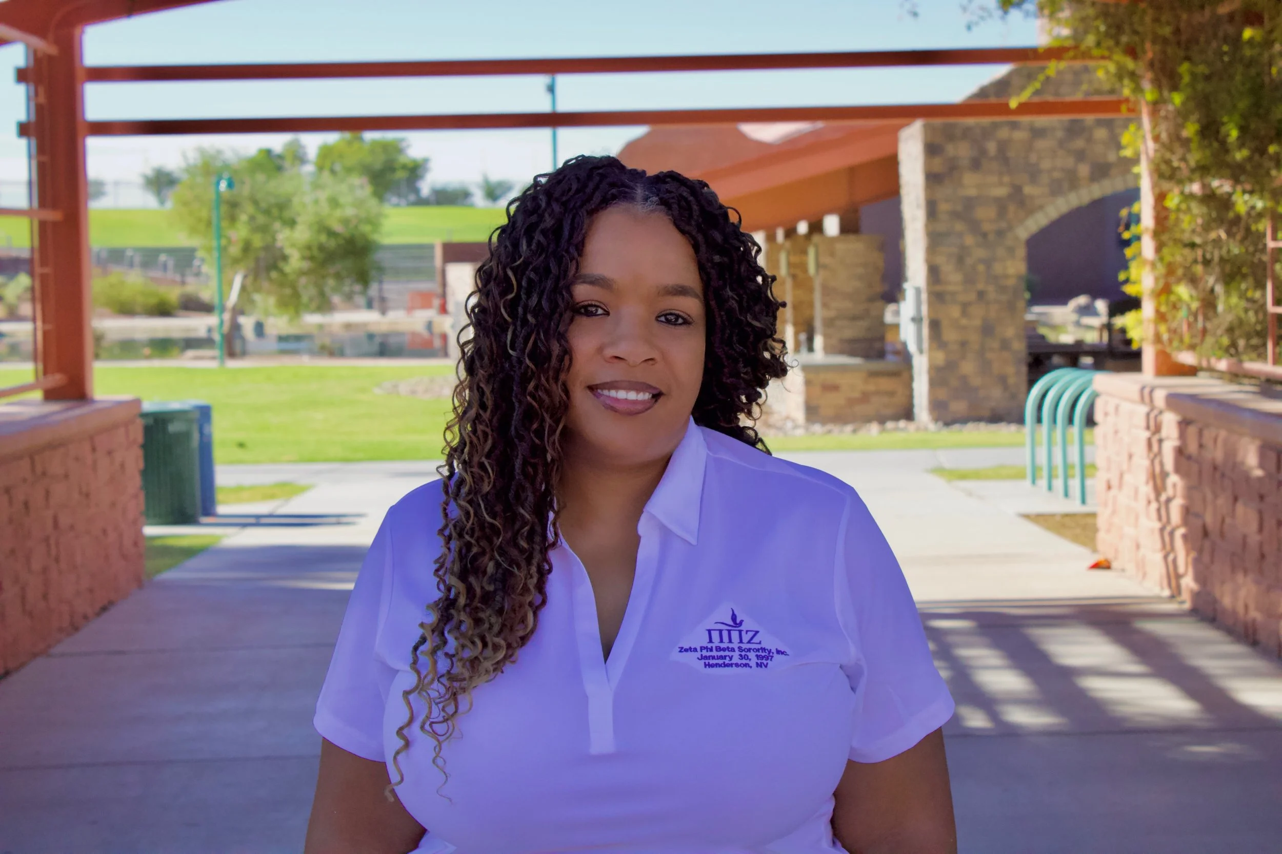 A woman with curly hair and a white polo shirt standing outdoors in a park with trees, grass, and a brick shelter in the background.