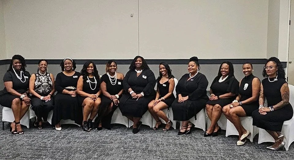 Group of eleven women sitting on chairs in a conference room, dressed in black formal attire with jewelry, smiling at the camera.