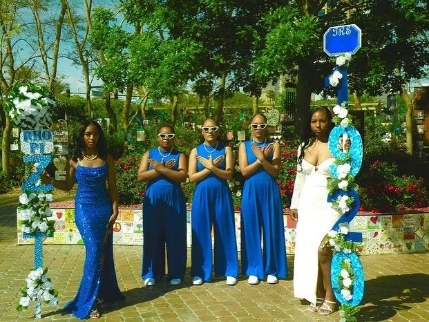Five young women standing outdoors in front of a garden with trees and flowers, dressed in elegant blue and white dresses, one holding a decorated number 20 sign with flowers.