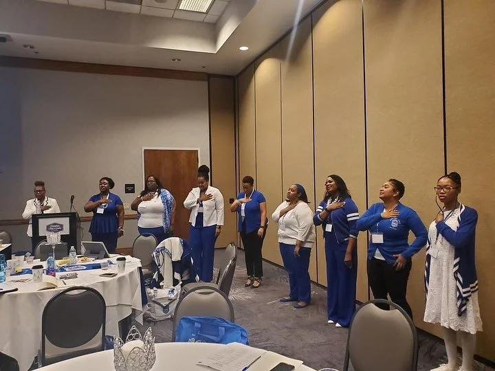 A group of women standing with their right hands over their hearts in a conference room, possibly during a pledge or national anthem.