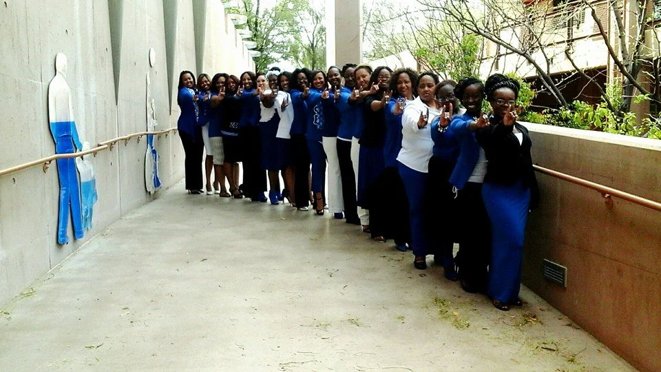 Group of women in blue and white clothing standing in a line on a balcony, pointing at the camera with their right hands.