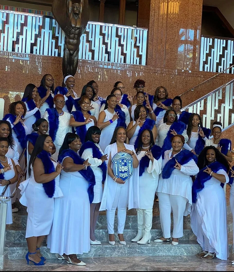 Group of women dressed in white with blue sashes and accessories, standing on a staircase in a large indoor space with a bronze statue of an angel or a saint behind them.