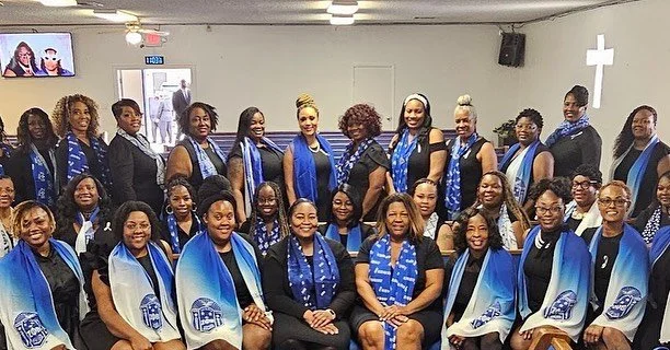 Group of women in church, dressed in black and blue, standing and sitting in a church hall, with a cross on the wall behind them.