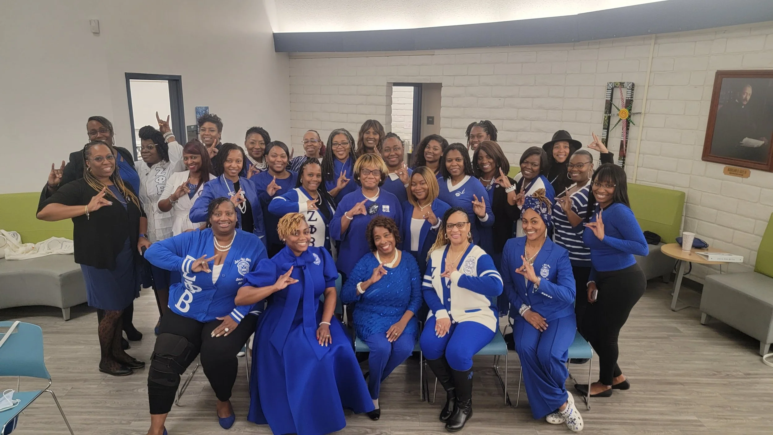 Group of women dressed in blue and black clothing, some wearing sorority or organization shirts, posing together in a room with white brick walls and green seating.