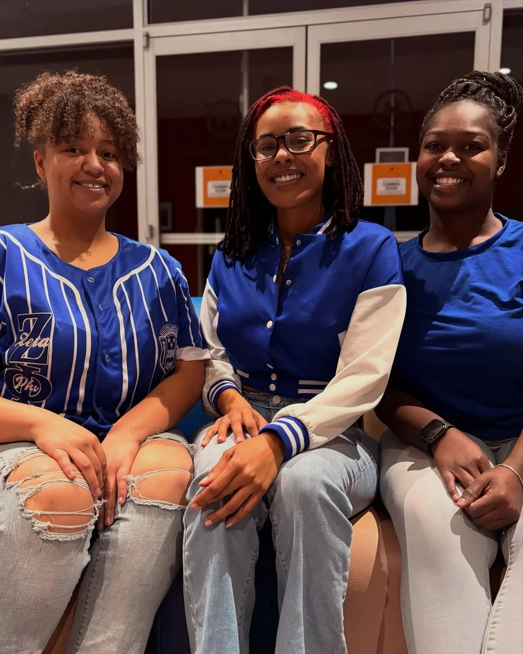 Three women sitting together in a casual setting, smiling at the camera, all wearing blue tops, two with denim jeans and one with white pants.