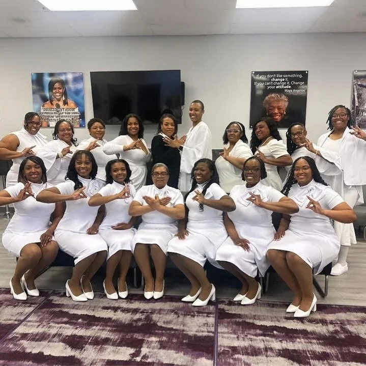 Group of women in white dresses and heels posing together in a room, some standing and some sitting, with motivational posters on the wall behind them.