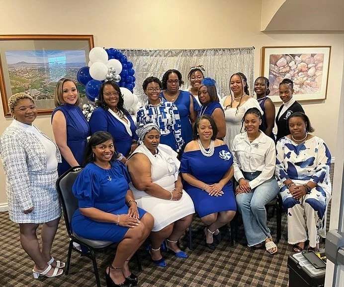 Group of women in blue and white outfits gathered in a room with framed artwork and balloons.