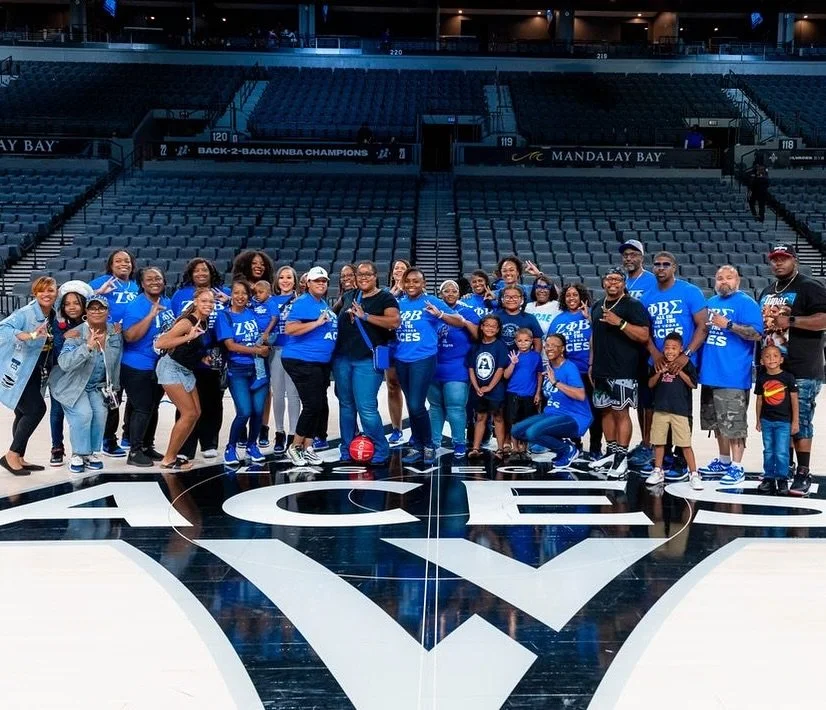 Group of people, including children and adults, posing for a photo on a basketball court inside an arena, with empty seats in the background. Many are wearing blue shirts, some with Greek letters, indicating a sorority or fraternity affiliation.