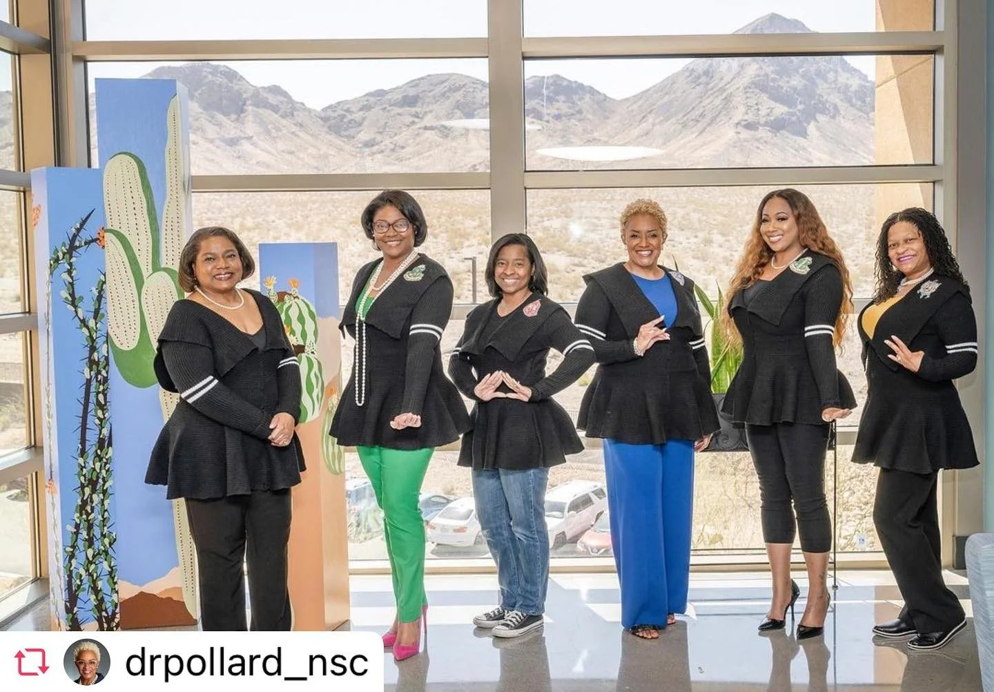 Six women standing indoors in front of a large window with mountain landscape views, dressed in coordinated black outfits with peplum jackets, some making hand gestures and smiling, with desert-themed paintings on display to the left.