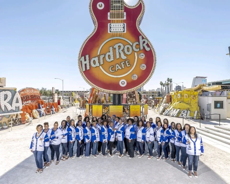 A group of women wearing blue and white jackets posing in front of the Hard Rock Cafe sign at the Neon Museum in Las Vegas with neon signs in the background.
